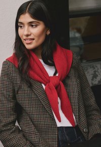 Sciarpa rossa lavorata a maglia, annodata al collo, sovrapposta a un blazer marrone a motivo pied de poule, indossata con una camicia bianca e denim. Tessuto liscio, colori a contrasto.