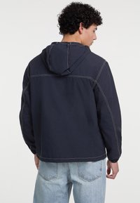 Giacca a cappuccio blu navy con cuciture bianche a contrasto, leggero design a spalla scesa e vestibilità rilassata. Il tessuto appare liscio e resistente.