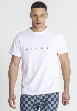 Mand iført hvid t-shirt med teksten "GIANNI" og blå ternede shorts, der står mod en ensfarvet baggrund.