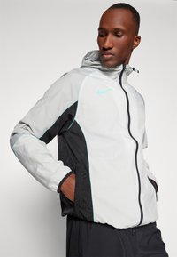 Veste légère à capuche grise et noire avec des accents turquoise, dotée d'une fermeture éclair et de poches latérales. Matériau résistant au vent.