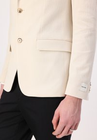 Crème kleurige textuur blazer met twee knopen, een borstzak en een zichtbare Calvin Klein label op de mouw. Gecombineerd met zwarte pantalon.