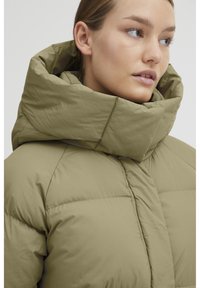 Doudoune dans un vert olive atténué, avec un col haut, un design matelassé et une texture lisse. Fermée par une fermeture éclair à l'avant, avec des coutures minimales.