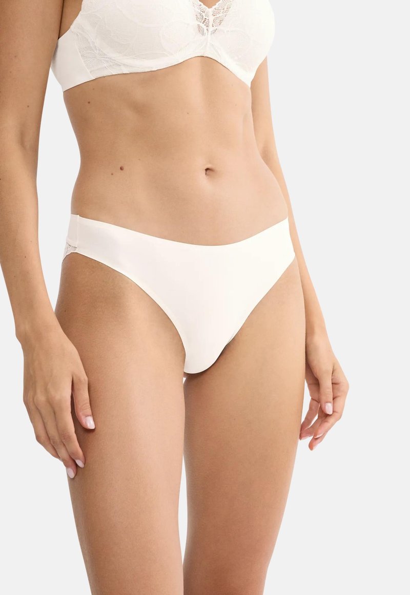 Γυναίκα που φοράει λευκό δαντελένιο σουτιέν και λευκό μαλακό εσώρουχο στυλ μπικίνι, στέκεται μπροστά σε απλό φόντο.