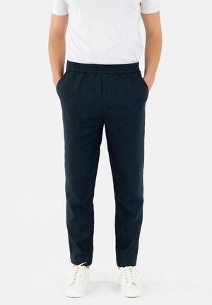 Mann iført marineblå bukser med elastisk midje, hvit t-skjorte og hvite joggesko, står med hendene i lommer.