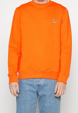 Personne portant un sweat-shirt à manches longues orange vif avec un petit logo zèbre sur la poitrine gauche, associé à un jean bleu clair.