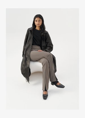 Czarny skórzany płaszcz, czarna koszula, beżowe spodnie z wysokim stanem, czarne buty na kwadratowym nosku ze srebrną klamrą, siedzący na białym krześle.