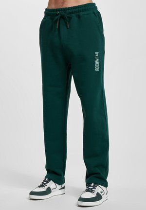 Grønne sweatpants med snor i taljen, ribbede manchetter og "ROCAWEAR" logo trykt i hvid på venstre lår. Sammen med hvide sneakers.