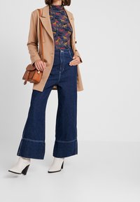 Beige skräddarsydd kappa över en marinblå topp med färgglada hästmönster, i kombination med vida mörkblå jeans och vita klackboots. Brun väska.