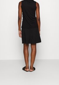 Person som står med ryggen mot kameran i en ärmlös svart topp, svart kjol och svarta platta sandaler mot en enfärgad bakgrund.