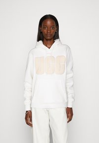 Biela mikina s kapucňou s výrazným béžovým logom "UGG" na prednej strane, s priestrannou kapsou a nastaviteľnou kapucňou.