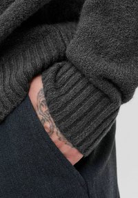 Mørkegrå strikket sweater med ribbet manchet, der delvist dækker en hånd, som er stukket ind i en lomme på teksturerede marineblå bukser.