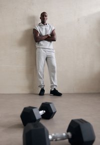 Pelēks sporta krekls un bikses ar elastīgām aprocēm, uz melnajām sporta kurpēm, stāvot blakus melniem sešstūra hantelēm uz gludas grīdas.