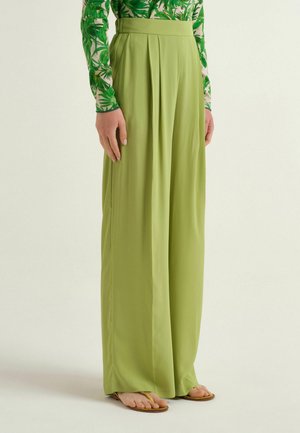 Femme portant un pantalon vert clair taille haute à jambes larges avec des plis, associé à un haut à manches longues à motif de feuilles tropicales et des sandales.