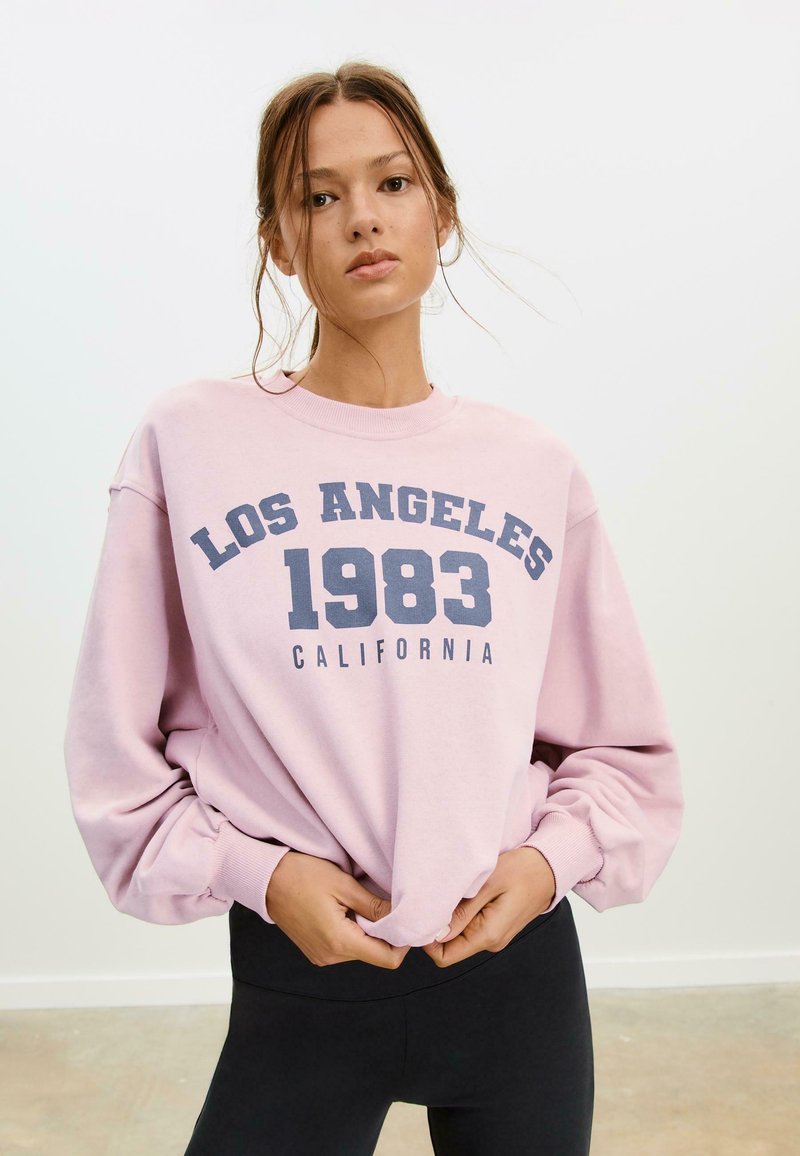 Ružová mikina s potlačou "LOS ANGELES 1983 CALIFORNIA" v námorníckej modrej. Dlhé rukávy s rebrovanými manžetami, voľný strih. Čierne legíny.