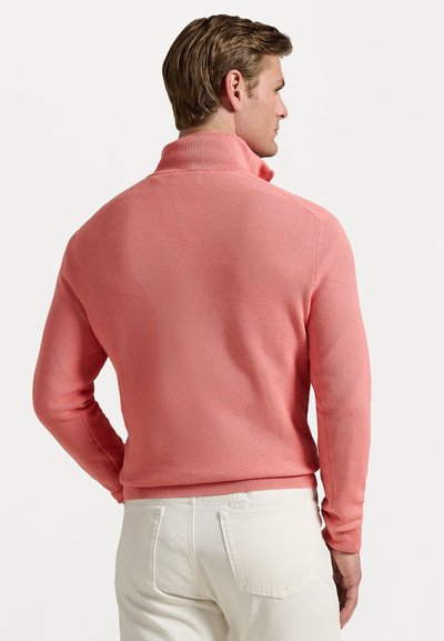 Pull-over en maille corail avec un col montant, des poignets et un ourlet côtelés ; motif texturé ; porté avec un pantalon blanc. Vue de dos.