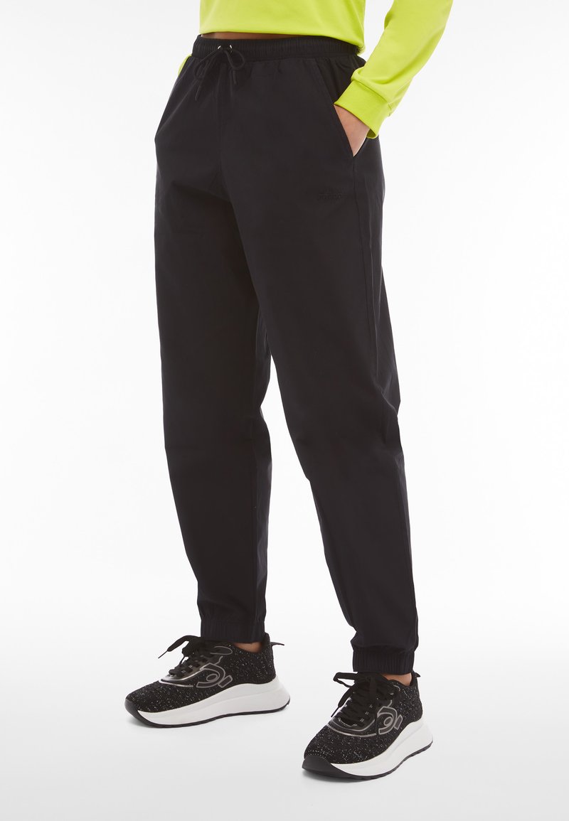 Schwarze Jogginghosen mit elastischem Bund, Seitentaschen und elastischen Bündchen. Kombiniert mit einem leuchtend grünen Langarmoberteil und schwarzen Turnschuhen.