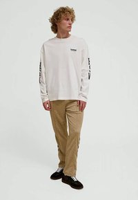 Giovane uomo con capelli ricci in piedi, che indossa una camicia bianca a maniche lunghe con scritte sulle maniche, pantaloni beige e sneakers nere.
