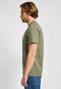 Oliwkowa koszulka bawełniana z krótkim rękawem, okrągłym dekoltem i małą naszywką graficzną na piersi. Slim fit i miękka tekstura.