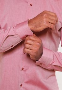 Chemise rose clair à manches longues avec poignets boutonnés. Présente une texture subtile, des boutons marron et une coupe ajustée. Aucun motif visible.