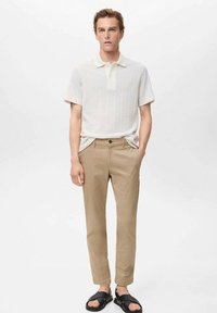 Polo shirt bianco a maniche corte con texture a coste, abbinato a pantaloni khaki slim-fit e sandali neri, in piedi su uno sfondo semplice.