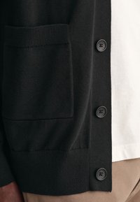 Schwarze Strickjacke aus Textilgewebe mit einer strukturierten Masche, die eine Fronttasche, fünf schwarze Knöpfe sowie einen gerippten Saum und Ärmel hat. Ein weißes Unterhemd ist sichtbar.
