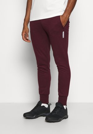 Hombre con pantalones jogger burdeos y zapatillas negras, de pie contra un fondo blanco, con una mano en el bolsillo.