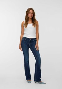 Moteris su ilgais plaukais stovi vilkėdama baltą be rankovių marškinėlį, tamsiai mėlynus bootcut džinsus ir šviesiai mėlynas smailianos papėdžiu batus prieš paprastą foną.
