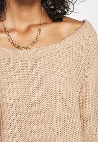 Pull beige à épaules dénudées avec un design en tricot ouvert. Il présente un large décolleté et des poignets côtelés. Accompagné d'un collier en chaîne dorée.