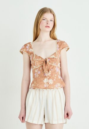 Bruine bloemrijke blouse met een vierkante halslijn, gestrikte voorkant, korte cap sleeves en kantafwerking. Gecombineerd met witte gestreepte shorts.