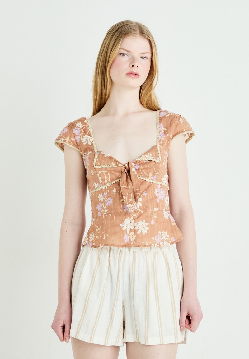 Brun blomstret bluse med firkantet utringning, bundet foran, korte kappeærmer og blondekant. Sammen med hvite stripete shorts.