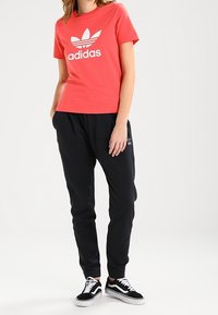Červené bavlněné tričko s bílým logem Adidas, spárované s černými sportovními kalhotami a černobílými teniskami. Neošizený, ležérní sportovní outfit.