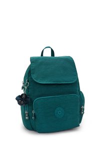Mochila de nailon color azul verdoso con una solapa, un bolsillo frontal con cremallera y un llavero de mono peludo. Presenta un distintivo con el logo en el bolsillo inferior.