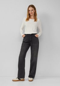 Jeans neri a gamba larga con una texture sbiadita, abbinati a una camicia bianca a maniche lunghe. La modella indossa sneakers in tonalità neutre, con le mani nelle tasche.