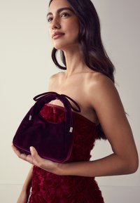 Geantă din catifea, de culoare vișinie închisă, cu formă rotunjită și mânere duble cu noduri. Dispune de accesorii argintii și textură moale.