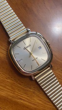 Montre Casio avec un cadran carré argenté, des aiguilles dorées, un guichet de date et un bracelet en maillons métalliques. Présente une finition polie et des index horaires minimalistes.