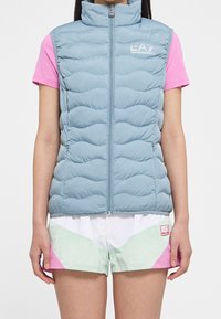 Gilet imbottito azzurro chiaro con colletto alto, caratterizzato da dettagli trapuntati e una zip frontale. Indossato sopra una camicia rosa e pantaloncini con accenti menta.