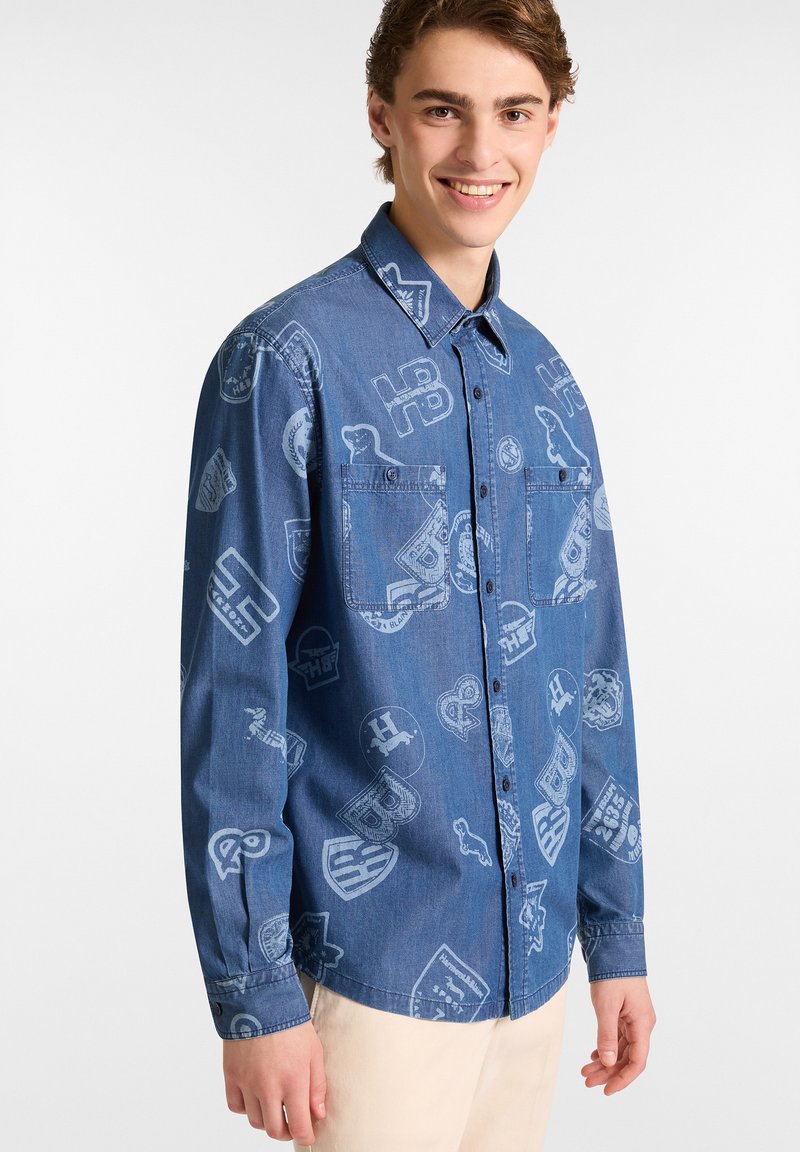 Blauwe denimshirt met een subtiel patroon van verschillende emblemen en logo's. Het heeft een knoopsluiting aan de voorkant en twee borstzakken. Langsleeves ontwerp.