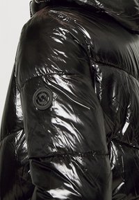 Chaqueta de plumas negra con textura brillante y acolchada, y un acento de logo circular en la manga. Capucha ajustable integrada en el diseño.