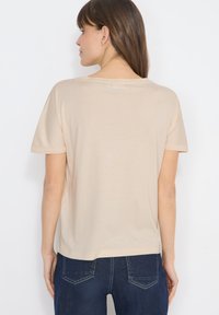 Femme aux cheveux bruns et raides, portant une chemise beige à manches courtes et un jean bleu foncé, debout face à un fond blanc, dos tourné vers le spectateur.