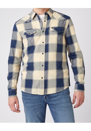 Mann trägt ein blau-creme-grau kariertes Hemd mit Klappentaschen und Blaumann, sichtbar von Hals bis Hüfte.