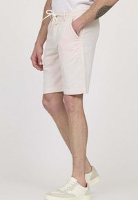 Shorts décontractés rose clair en tissu doux, dotés de poches latérales et d'une taille élastique avec cordon de serrage. Portés avec des baskets beiges.