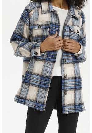 Woman wearing a blue, beige, and black plaid jacket over a white top and black pants, holding the jacket lapels with both hands.