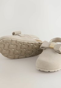 Des chaussures pour tout-petits beiges présentent un nœud décoratif et un accent doré, montrant une semelle texturée pour une adhérence sur un fond clair.