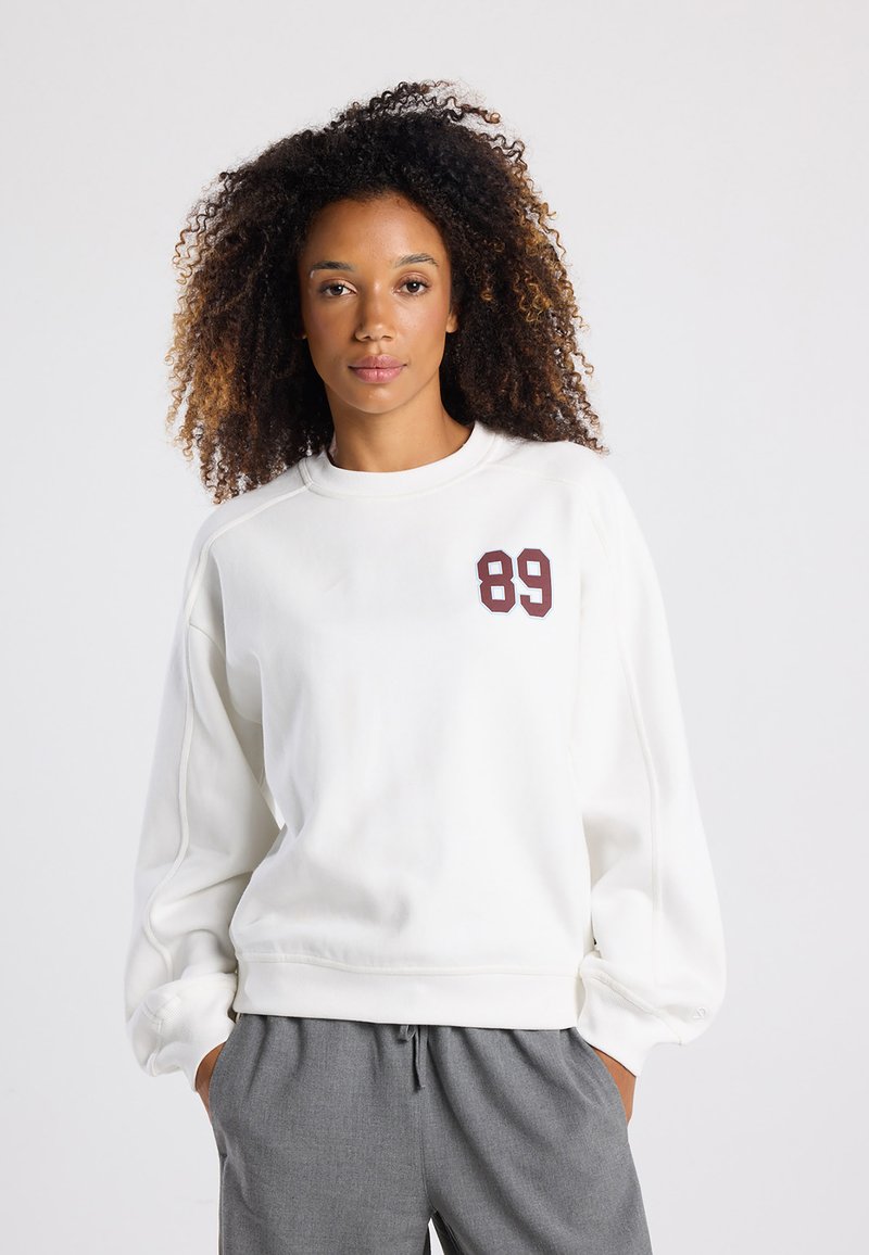 White sweatshirt with long sleeves, featuring a maroon "89" emblem on the left chest. Smooth fabric and relaxed fit. Worn with gray trousers.
