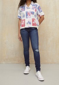Camicia a maniche corte in tessuto leggero con bottoni, decorata con motivi floreali in blu, rosso e rosa, abbinata a jeans skinny blu scuro e scarpe da ginnastica bianche.