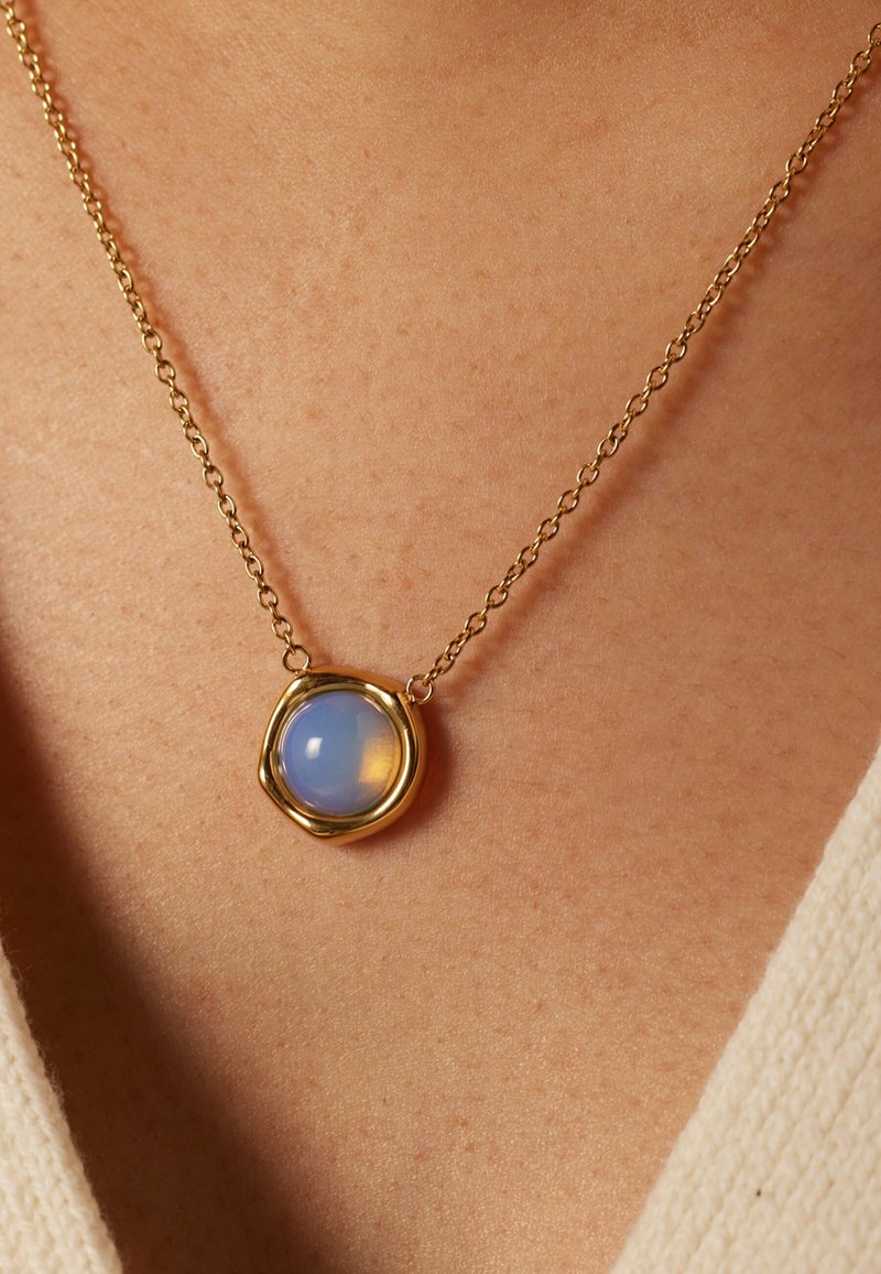 Gouden ketting met een ronde, transparante opalescente hanger met een textuur bezel, rustend tegen een textuur huidoppervlak.