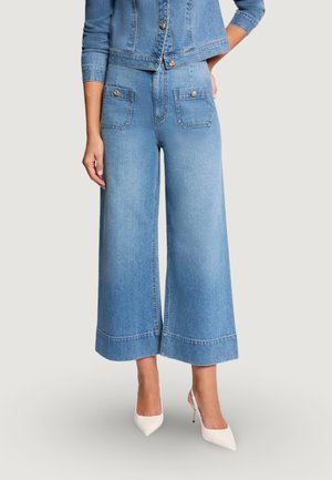 Mujer con vaqueros acampanados de pierna ancha y corte cropped en azul claro, con bolsillos delanteros con botones, combinados con chaqueta vaquera a juego y tacones blancos puntiagudos.