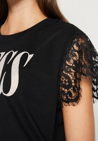 Camiseta negra de manga corta con borde de encaje negro, adornada con letras de strass plateadas en la parte delantera. Tejido de algodón suave.