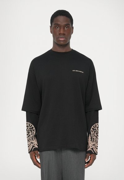 Giovane in camicia nera a maniche lunghe con la scritta "LES BENJAMINS" e disegni beige a motivi sulle maniche, abbinata a pantaloni grigi.