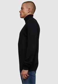 Svart genser med høy hals og lange ermer, ribbestrikket krage og myk tekstur, iført med blå jeans, vist fra siden.